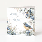 A square greeting card standing upright on a white surface against a white background. The card features a winter illustration with snow-covered branches, blue berries, and a small bird in blue, grey, cream, and red tones.