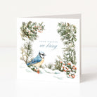 A square white Christmas greeting card featuring a watercolor illustration of a blue jay in a snowy winter scene, surrounded by green pine branches with small red berries and the text 'GOOD TIDINGS we bring'.