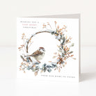 A square Christmas greeting card standing upright on a white surface. The card features a watercolour-style wreath with snow, leaves, berries, and a small bird, with the text 'Wishing you a very merry christmas' and 'From our home to yours'.