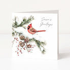 A white greeting card standing upright on a light neutral surface. The front features an illustration of a red cardinal perched on snowy pine branches with pinecones, and the text 'Season's Greetings' in black script.