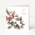A white Christmas greeting card standing upright on a light surface, printed with a red cardinal perched on snowy evergreen branches with pinecones and the words 'Let It Snow' in black script.
