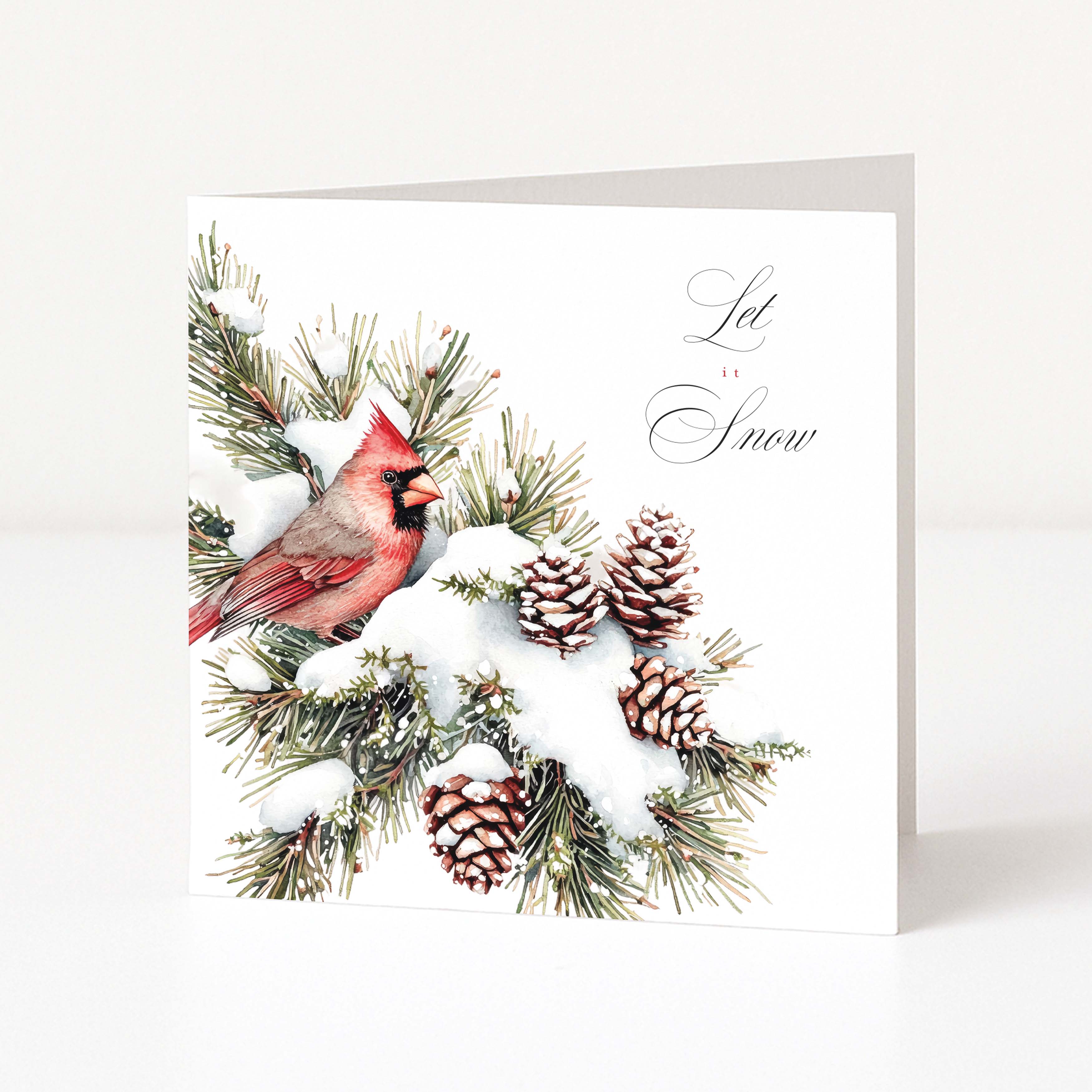 A white Christmas greeting card standing upright on a light surface, printed with a red cardinal perched on snowy evergreen branches with pinecones and the words 'Let It Snow' in black script.
