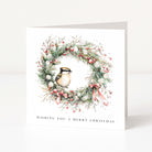 A square white Christmas greeting card standing upright on a light grey surface. The front features an illustrated wreath with green pine branches, red berries, white snow-like accents, and a small bird at the centre, with the text 'WISHING YOU A MERRY CHRISTMAS' printed below.
