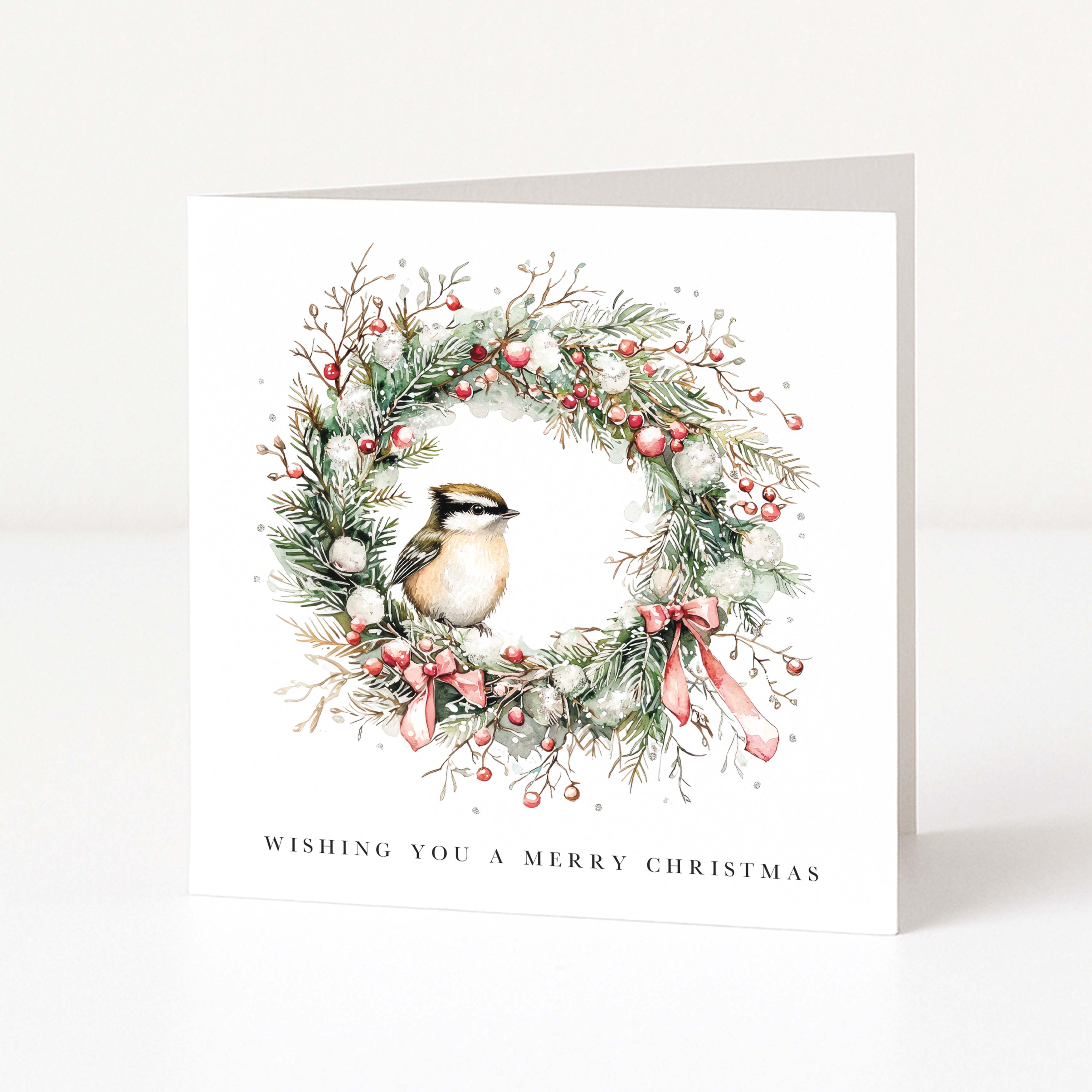 A square white Christmas greeting card standing upright on a light grey surface. The front features an illustrated wreath with green pine branches, red berries, white snow-like accents, and a small bird at the centre, with the text 'WISHING YOU A MERRY CHRISTMAS' printed below.