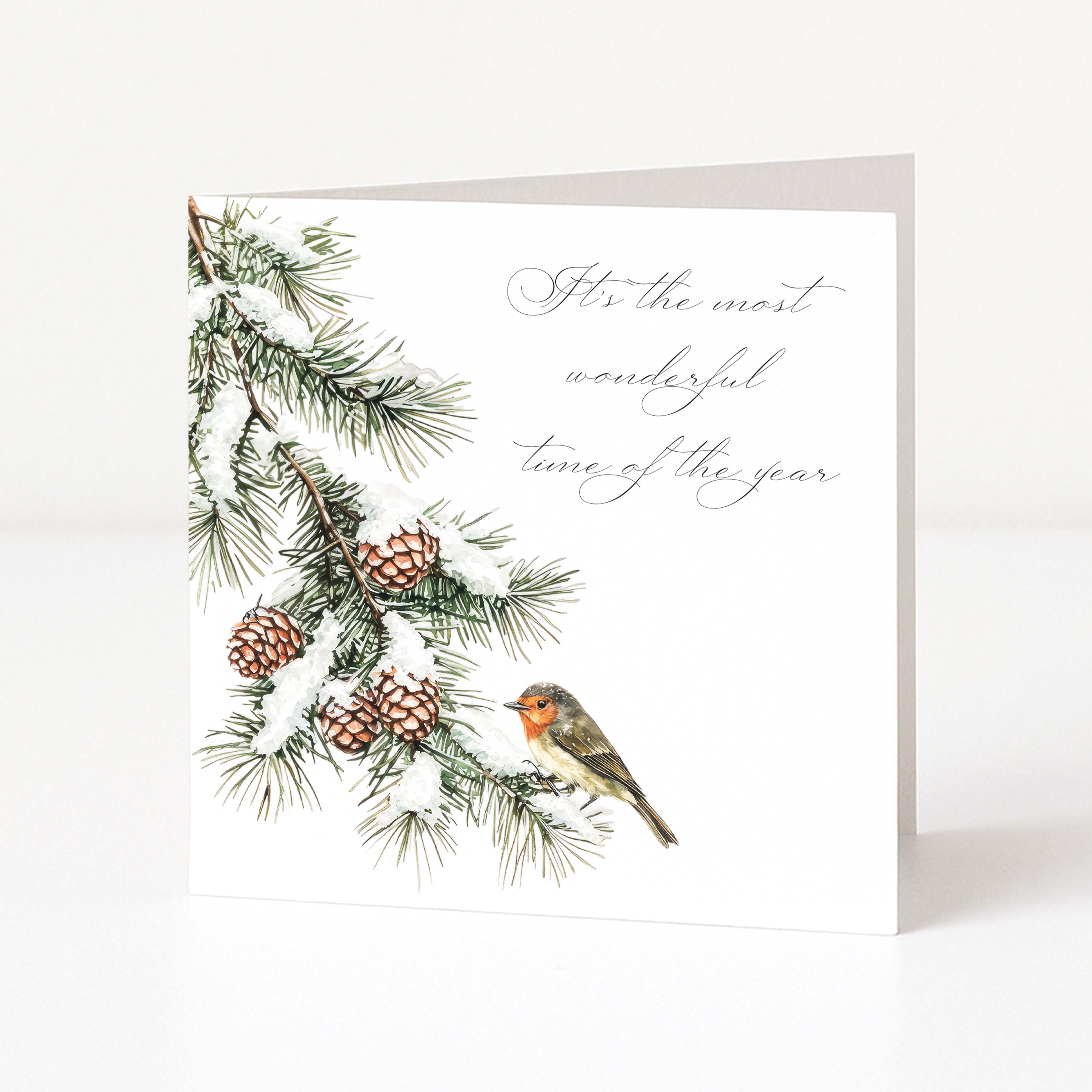 A square white greeting card standing upright, printed with a winter pine branch, pinecones, a small bird, and the phrase “It’s the most wonderful time of the year” in gray script.