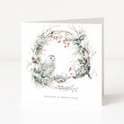 A square white Christmas greeting card standing upright on a white surface, featuring a watercolour-style winter wreath with a snowy owl, branches, and small red berries. The text 'SEASON'S GREETINGS' appears at the bottom.