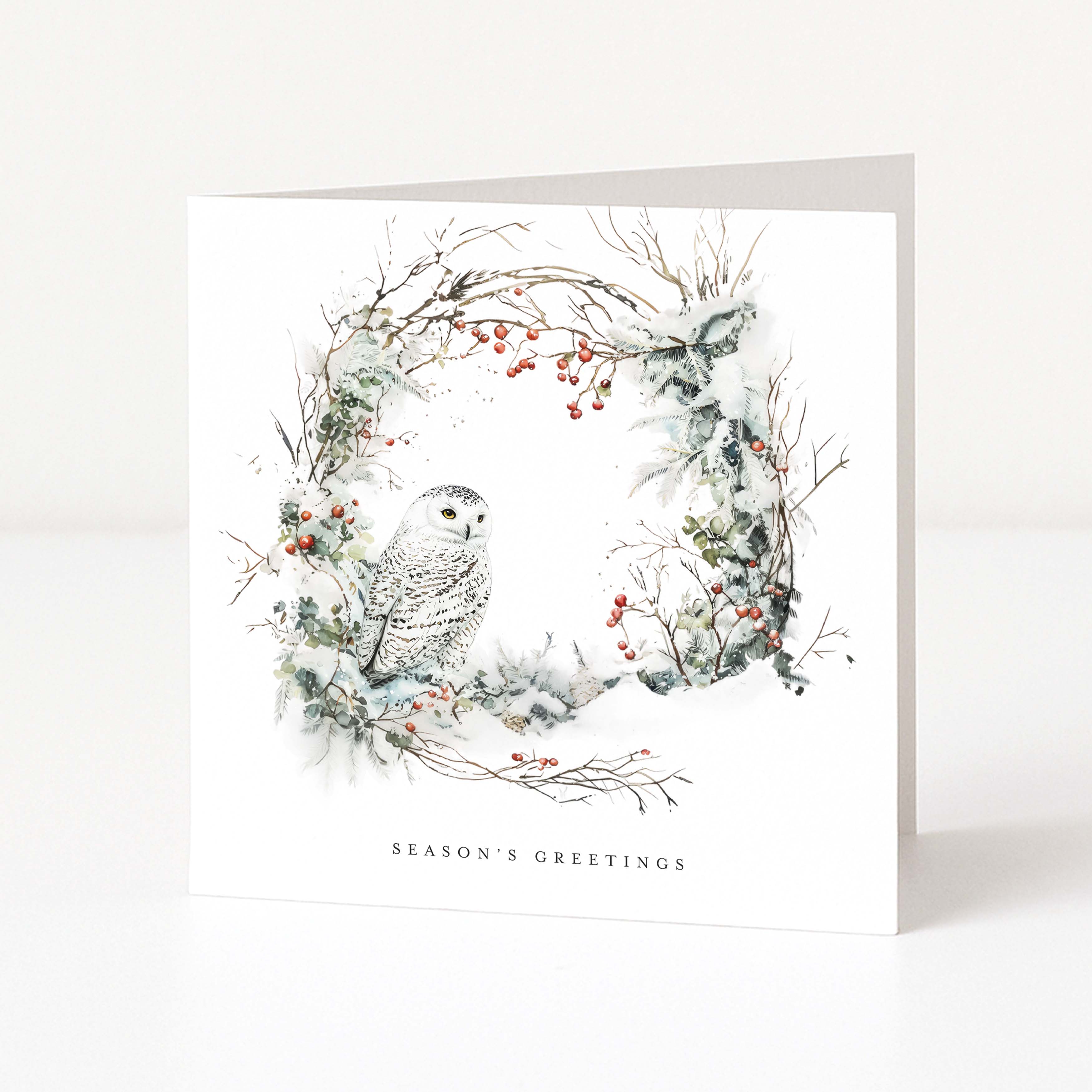A square white Christmas greeting card standing upright on a white surface, featuring a watercolour-style winter wreath with a snowy owl, branches, and small red berries. The text 'SEASON'S GREETINGS' appears at the bottom.