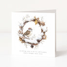 A square white greeting card standing upright on a light neutral surface. The front features an illustrated winter wreath with snow, brown leaves, pinecones, and a small bird, with the text 'wishing you a merry christmas and a happy new year' below.