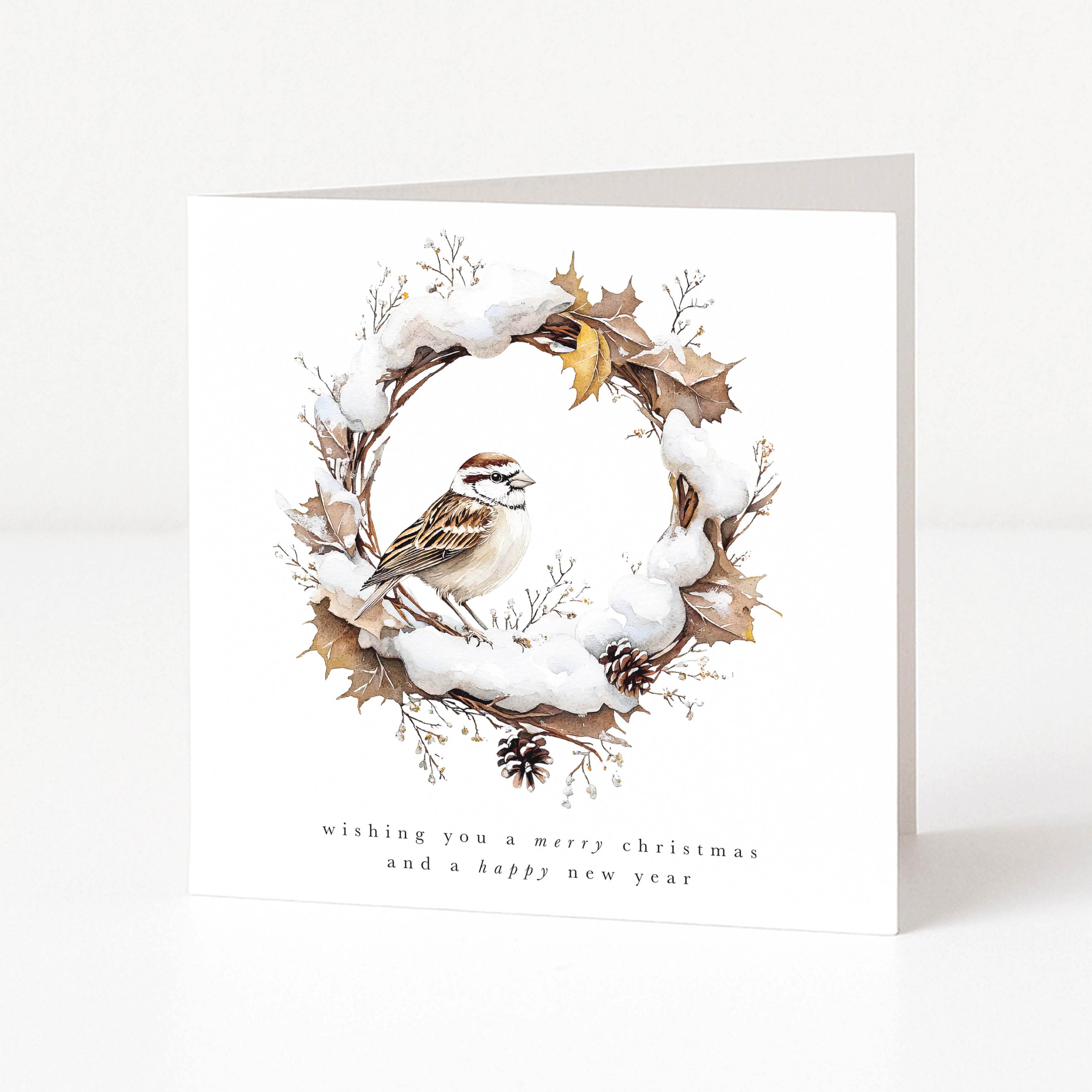 A square white greeting card standing upright on a light neutral surface. The front features an illustrated winter wreath with snow, brown leaves, pinecones, and a small bird, with the text 'wishing you a merry christmas and a happy new year' below.
