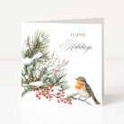 A square white holiday greeting card standing upright on a light neutral surface. The card features a watercolor-style winter illustration with snow-dusted pine branches, red berries, and a robin bird on the lower right, alongside the words “HAPPY Holidays” in red and gray script.