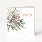 A square Christmas greeting card standing upright on a white surface. The card features an illustration of snowy evergreen branches with red berries and a small cardinal, with the text 'Happy Christmas' and 'from our home to yours'.