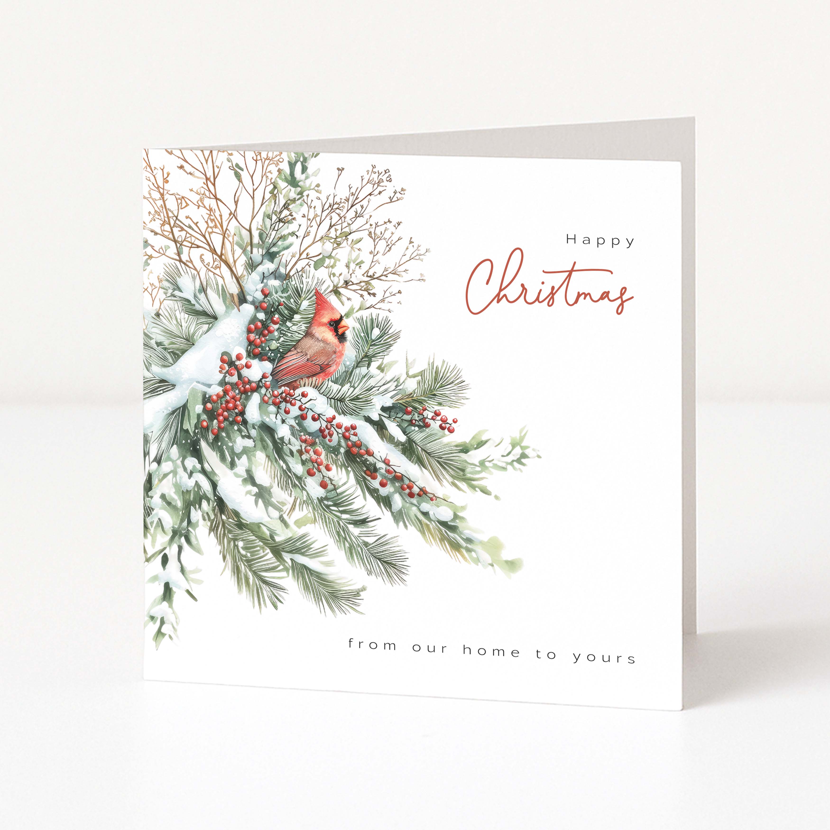 A square Christmas greeting card standing upright on a white surface. The card features an illustration of snowy evergreen branches with red berries and a small cardinal, with the text 'Happy Christmas' and 'from our home to yours'.