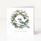 A white Christmas greeting card standing upright, featuring a watercolour illustration of a blue jay inside a pine wreath with snow and pinecones, and the words 'Joy to the World' in light blue script at the bottom.