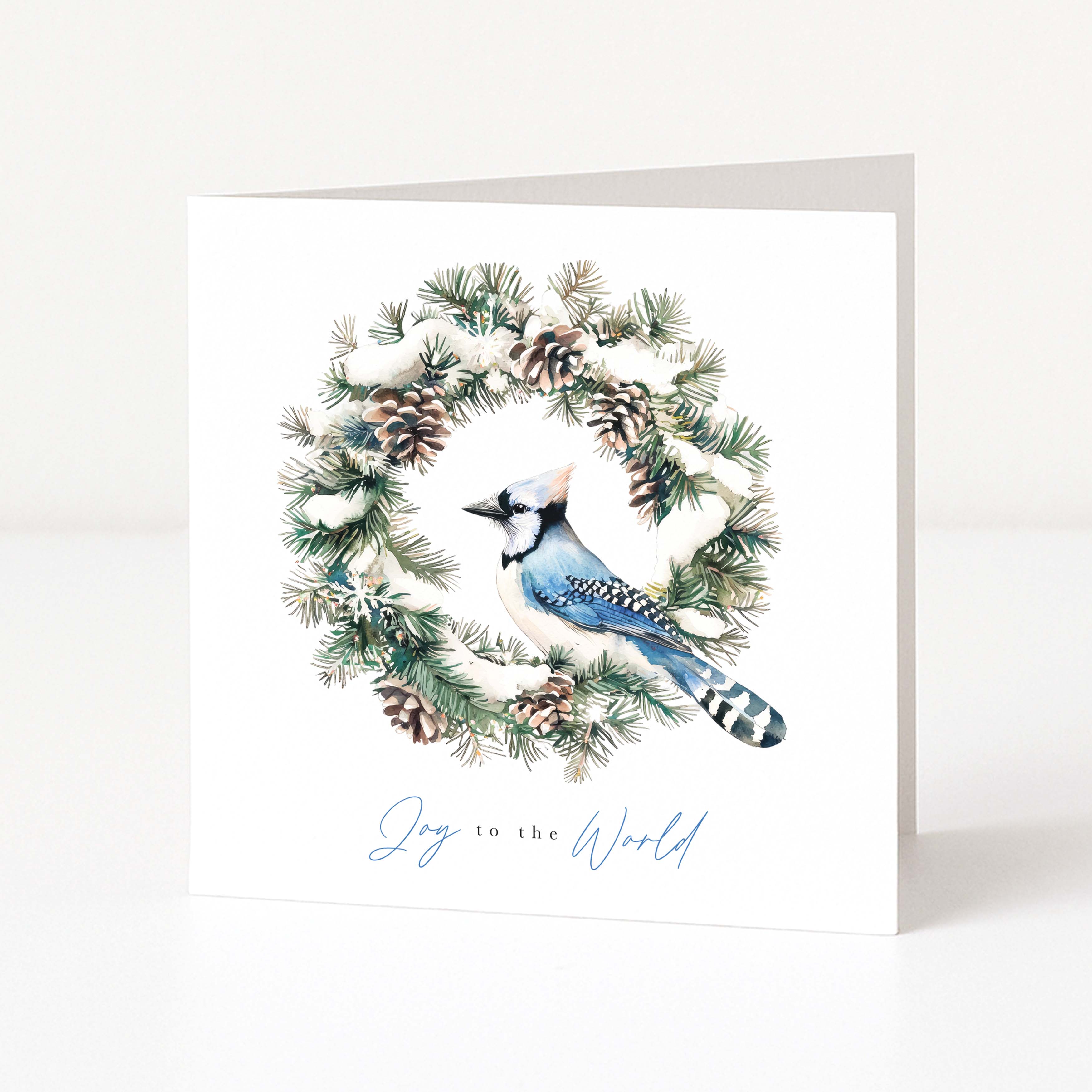 A white Christmas greeting card standing upright, featuring a watercolour illustration of a blue jay inside a pine wreath with snow and pinecones, and the words 'Joy to the World' in light blue script at the bottom.