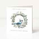 A square white Christmas greeting card standing upright on a light surface. The front features an illustration of a blue jay perched inside a snowy green wreath, with the text 'FROM OUR HOME TO YOURS' at the top and 'WE WISH YOU A MERRY CHRISTMAS' at the bottom.