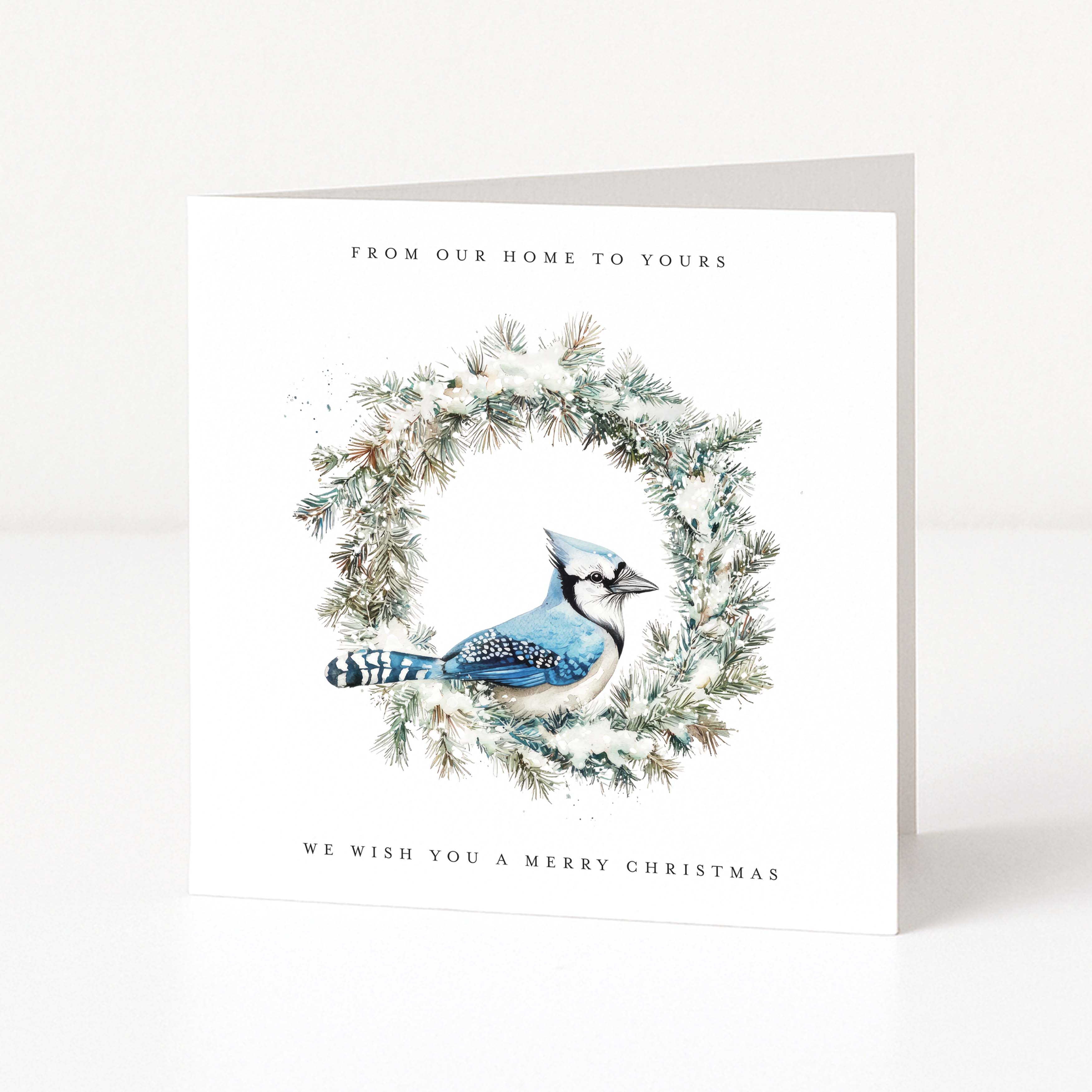 A square white Christmas greeting card standing upright on a light surface. The front features an illustration of a blue jay perched inside a snowy green wreath, with the text 'FROM OUR HOME TO YOURS' at the top and 'WE WISH YOU A MERRY CHRISTMAS' at the bottom.