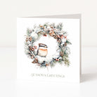 A white square Christmas greeting card standing upright on a light grey surface, featuring a winter wreath illustration with a small bird, pinecones, berries, and the text 'SEASON'S GREETINGS' at the bottom.
