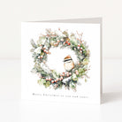 A square white Christmas greeting card standing upright on a light gray surface. The front features a watercolour-style wreath with green foliage, small red berries, and a brown-and-white bird at the center, with the text 'Merry Christmas to you and yours' printed below.