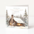 A square Christmas greeting card standing upright on a white surface. The card features a watercolor illustration of a snow-covered log cabin with warm glowing windows, surrounded by tall pine trees.