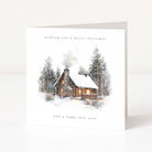 A square white Christmas greeting card standing upright on a light neutral surface. The front features an illustrated winter cabin with snow, evergreen trees, and soft smoke, with the text 'wishing you a merry christmas' at the top and 'and a happy new year' at the bottom.