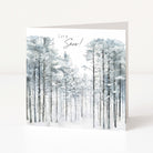 A square Christmas greeting card standing upright on a white surface, featuring a watercolour winter forest scene with tall snow-covered trees and the text 'Let it Snow!' at the top.