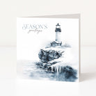 A square Christmas greeting card standing upright on a white surface, featuring a lighthouse scene with icy rocks and the text 'SEASON'S greetings' in blue-gray script and serif lettering.