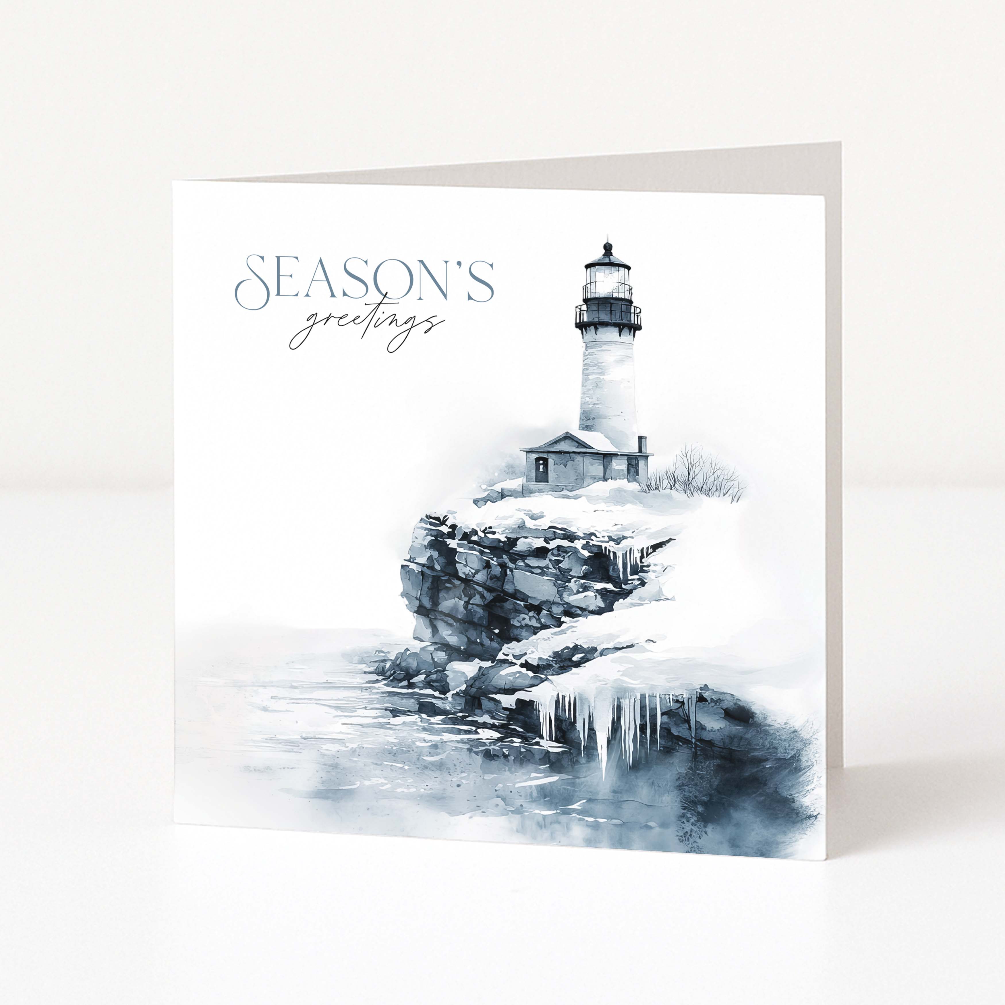 A square Christmas greeting card standing upright on a white surface, featuring a lighthouse scene with icy rocks and the text 'SEASON'S greetings' in blue-gray script and serif lettering.