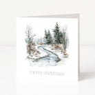 A white Christmas square greeting card standing upright on a white surface, featuring a watercolor winter landscape with a winding icy path, bare trees, evergreen trees, and the text 'merry christmas' at the bottom.