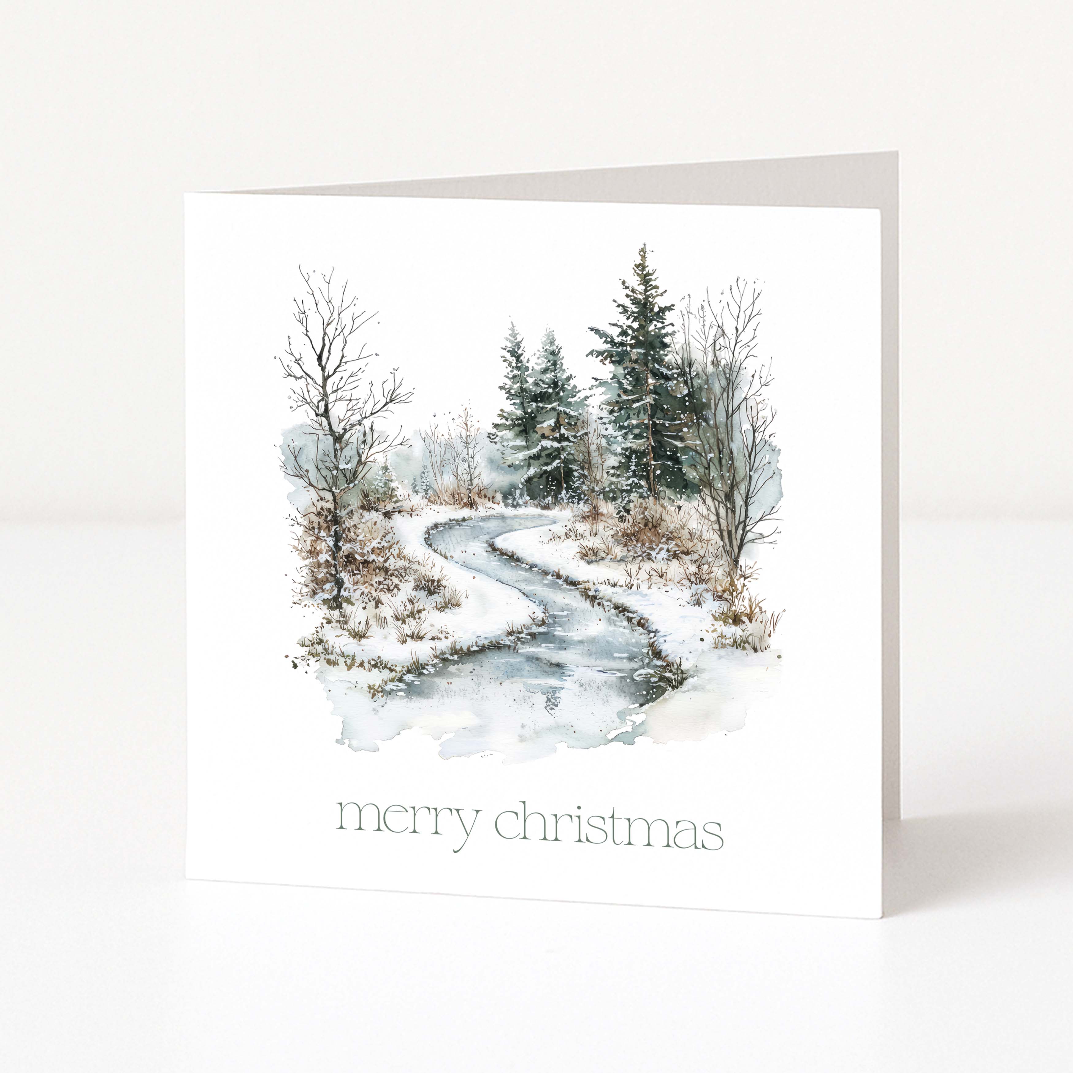 A white Christmas square greeting card standing upright on a white surface, featuring a watercolor winter landscape with a winding icy path, bare trees, evergreen trees, and the text 'merry christmas' at the bottom.