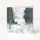 A square Christmas greeting card standing upright on a white surface, printed with a snowy forest path scene and a small white label reading 'wishing you a merry christmas'.