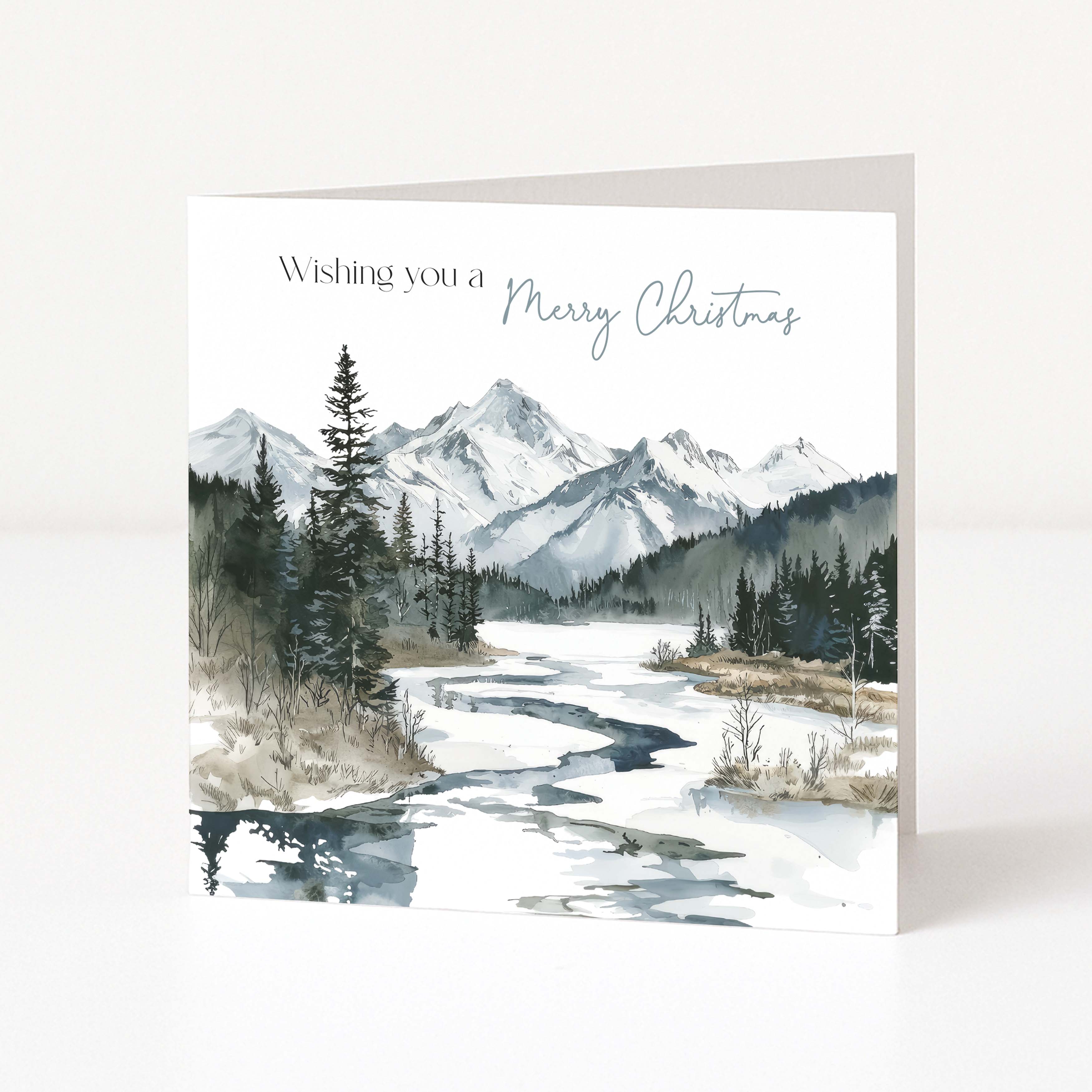 A square Christmas greeting card standing upright on a white surface. The front features a watercolor-style winter landscape with snowy mountains, evergreen trees, and a partially frozen river, with the text 'Wishing you a Merry Christmas' printed at the top.