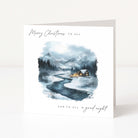 A square white Christmas greeting card standing upright on a light surface. The front features a watercolour winter landscape with mountains, a winding river, dark trees, and small glowing cabins.