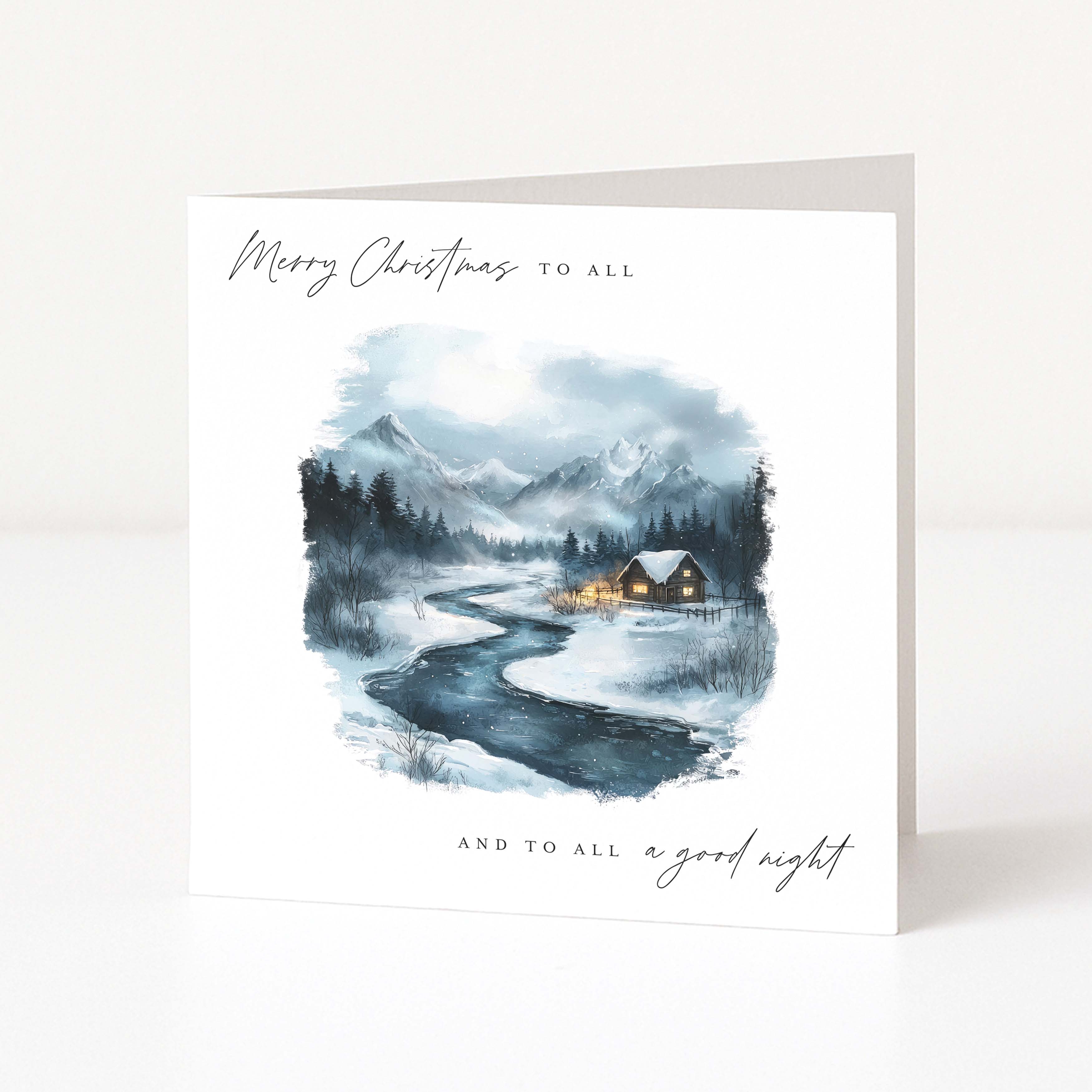A square white Christmas greeting card standing upright on a light surface. The front features a watercolour winter landscape with mountains, a winding river, dark trees, and small glowing cabins.