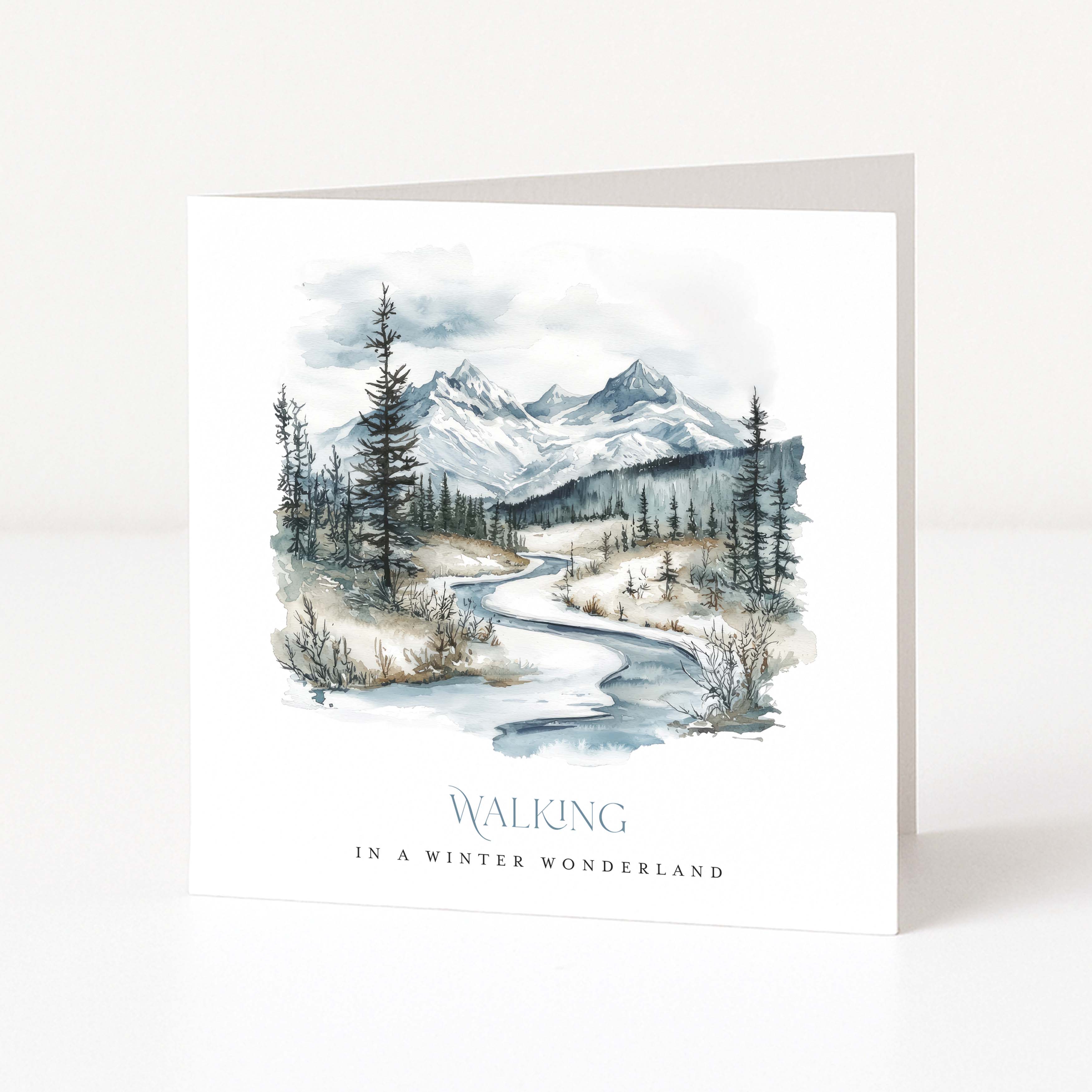 A white Christmas greeting card standing upright on a white surface, featuring a watercolor winter landscape with mountains, trees, and a winding river. The text at the bottom reads “WALKING IN A WINTER WONDERLAND.