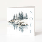 A white Christmas greeting card standing upright, printed with a winter landscape scene showing snow-covered rocks, evergreen trees, and their reflection in water, with the word 'NOEL' printed vertically in blue-gray letters on the right.