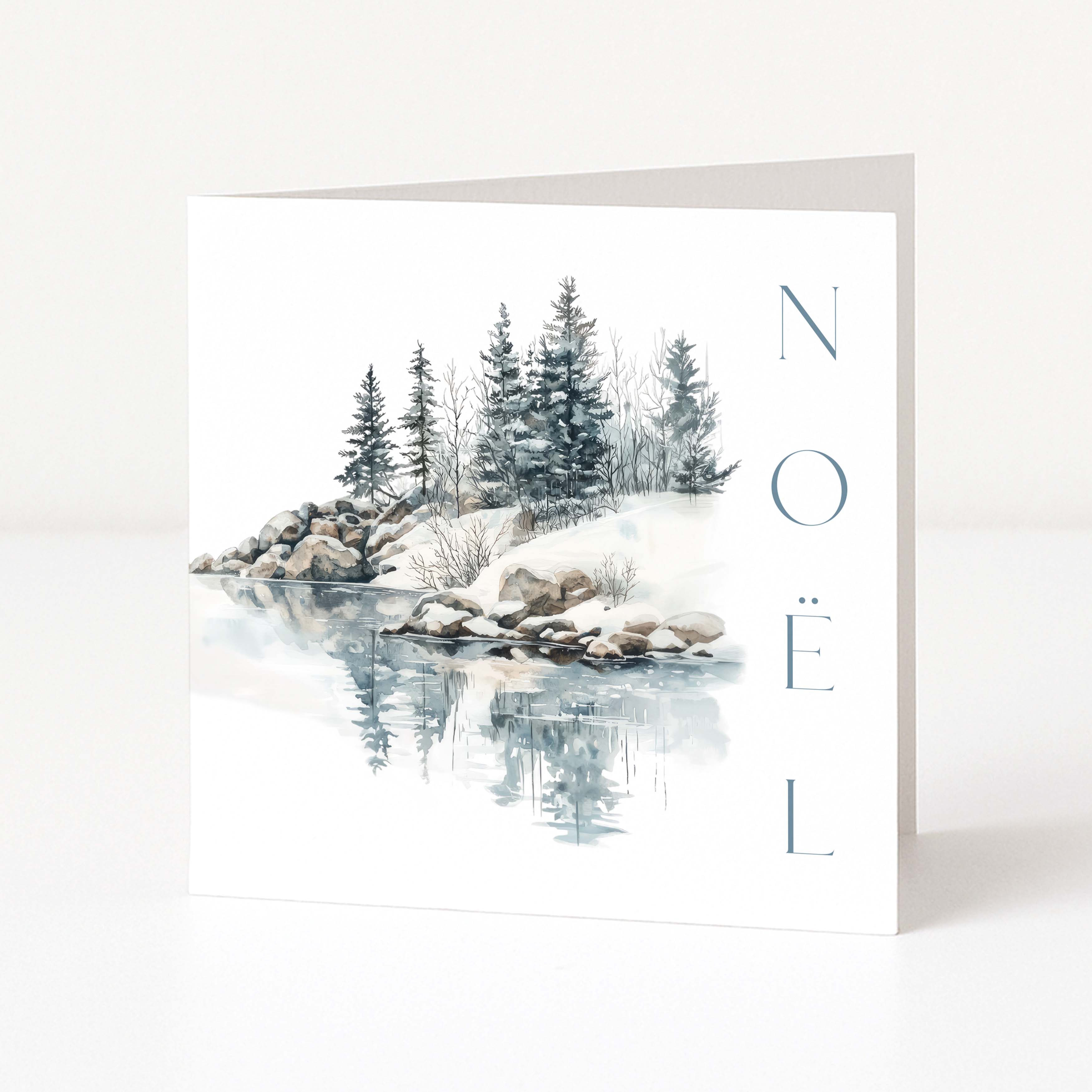 A white Christmas greeting card standing upright, printed with a winter landscape scene showing snow-covered rocks, evergreen trees, and their reflection in water, with the word 'NOEL' printed vertically in blue-gray letters on the right.