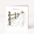 A square Christmas greeting card standing upright on a white surface, printed with a snowy landscape scene featuring a wooden fence, a footpath, and the words 'Let it snow' in blue script.