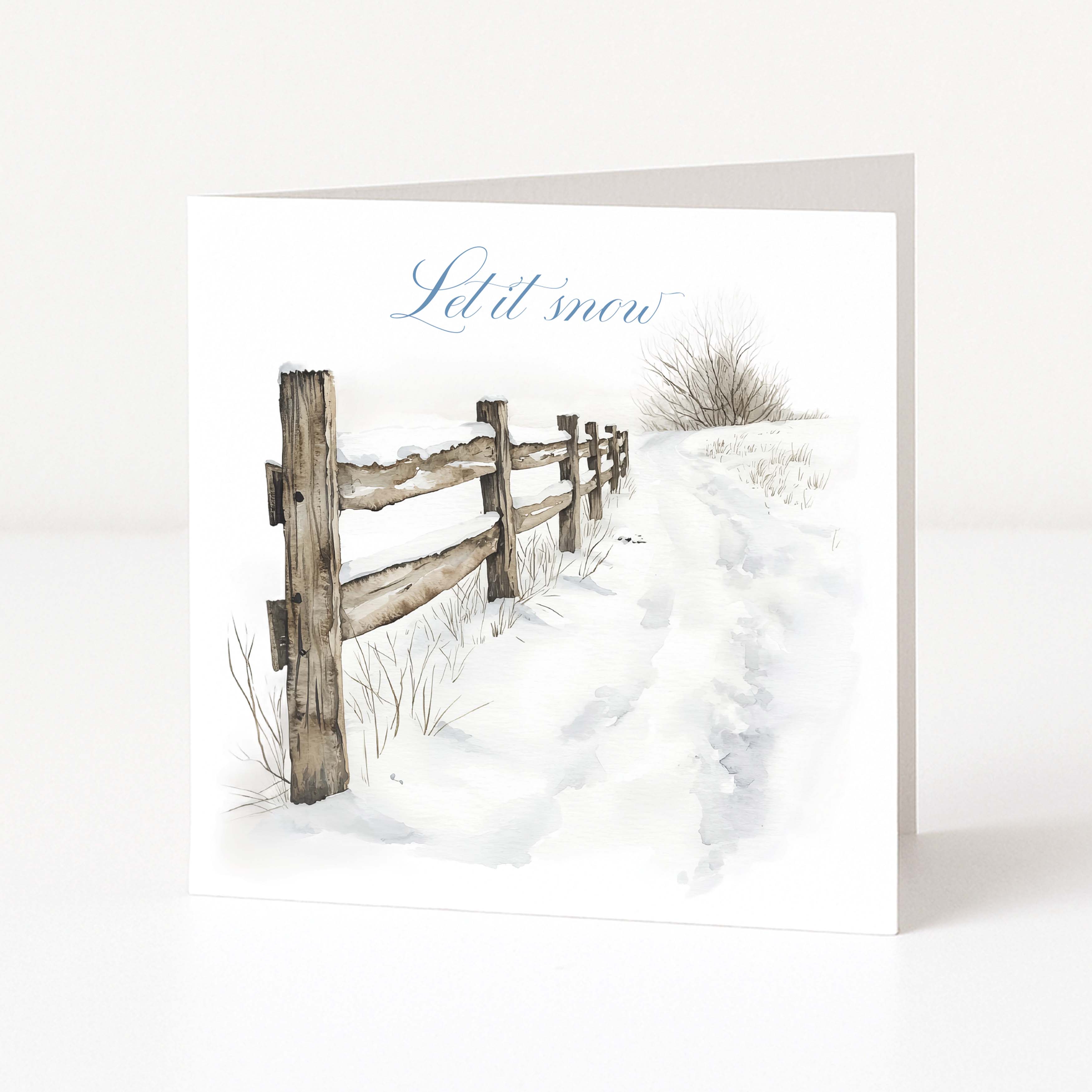 A square Christmas greeting card standing upright on a white surface, printed with a snowy landscape scene featuring a wooden fence, a footpath, and the words 'Let it snow' in blue script.