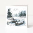 A square Christmas greeting card standing upright on a white surface. The front features a watercolor-style winter landscape with snow-covered ground, evergreen trees, and two small wooden boats in the foreground.