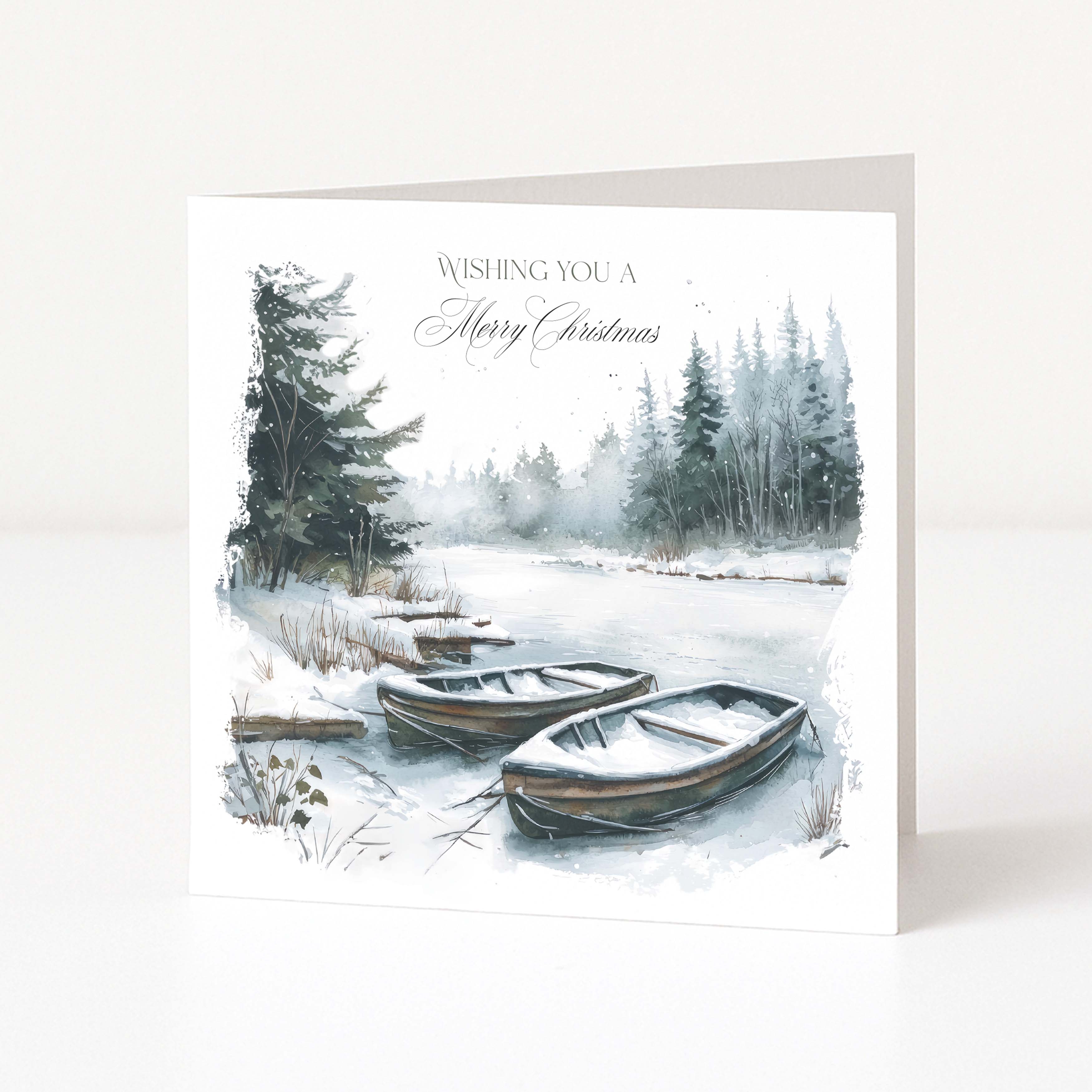 A square Christmas greeting card standing upright on a white surface. The front features a watercolor-style winter landscape with snow-covered ground, evergreen trees, and two small wooden boats in the foreground.
