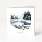 A white Christmas greeting card standing upright, printed with a winter lakeside scene featuring two rowboats, evergreen trees, and the words 'Happy Holidays' near the bottom.