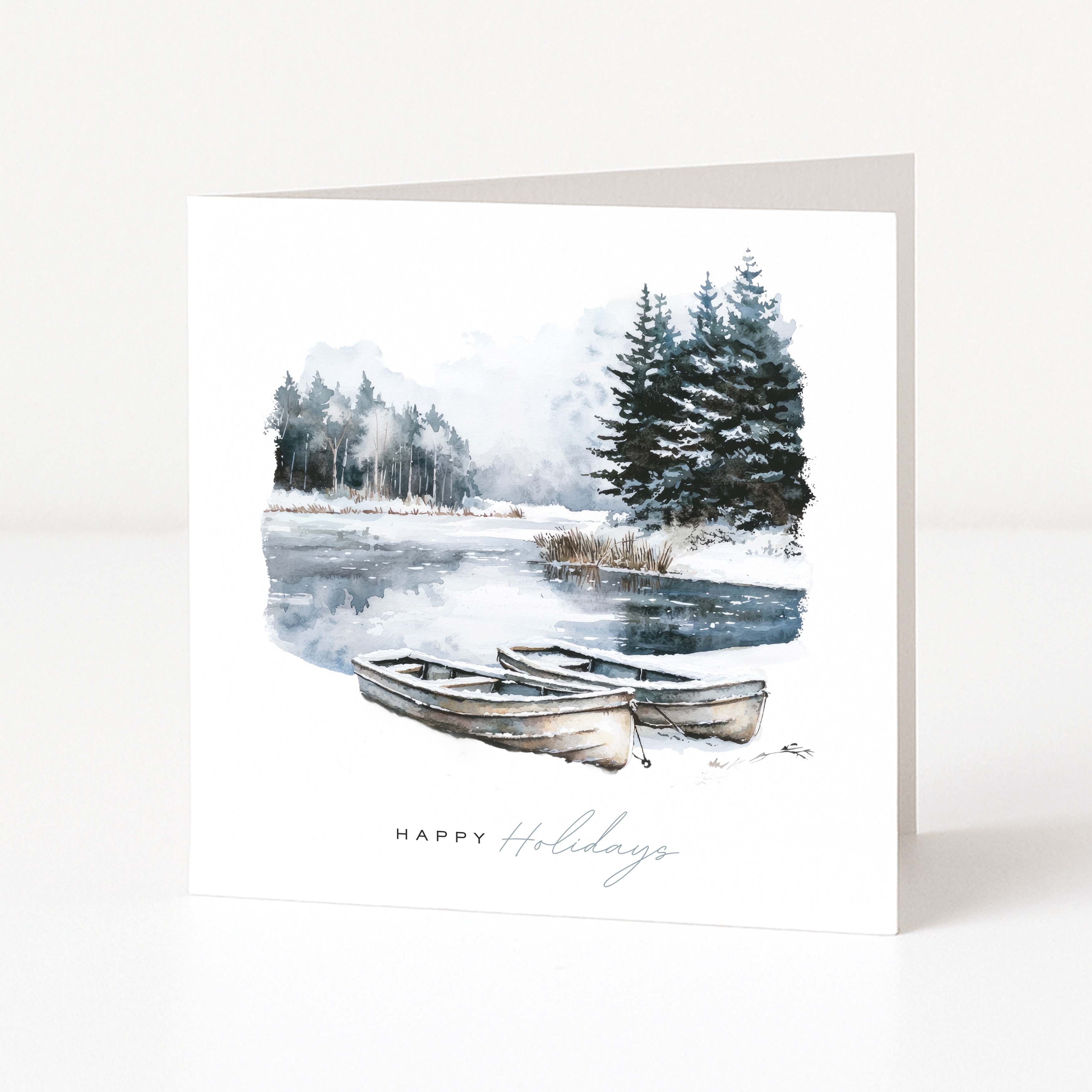 A white Christmas greeting card standing upright, printed with a winter lakeside scene featuring two rowboats, evergreen trees, and the words 'Happy Holidays' near the bottom.
