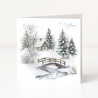 A square Christmas greeting card standing upright on a white surface, featuring a winter landscape illustration with snow, evergreen trees, a small cabin, a wooden bridge, and a stream. The card has the words 'let it snow' printed at the top right.