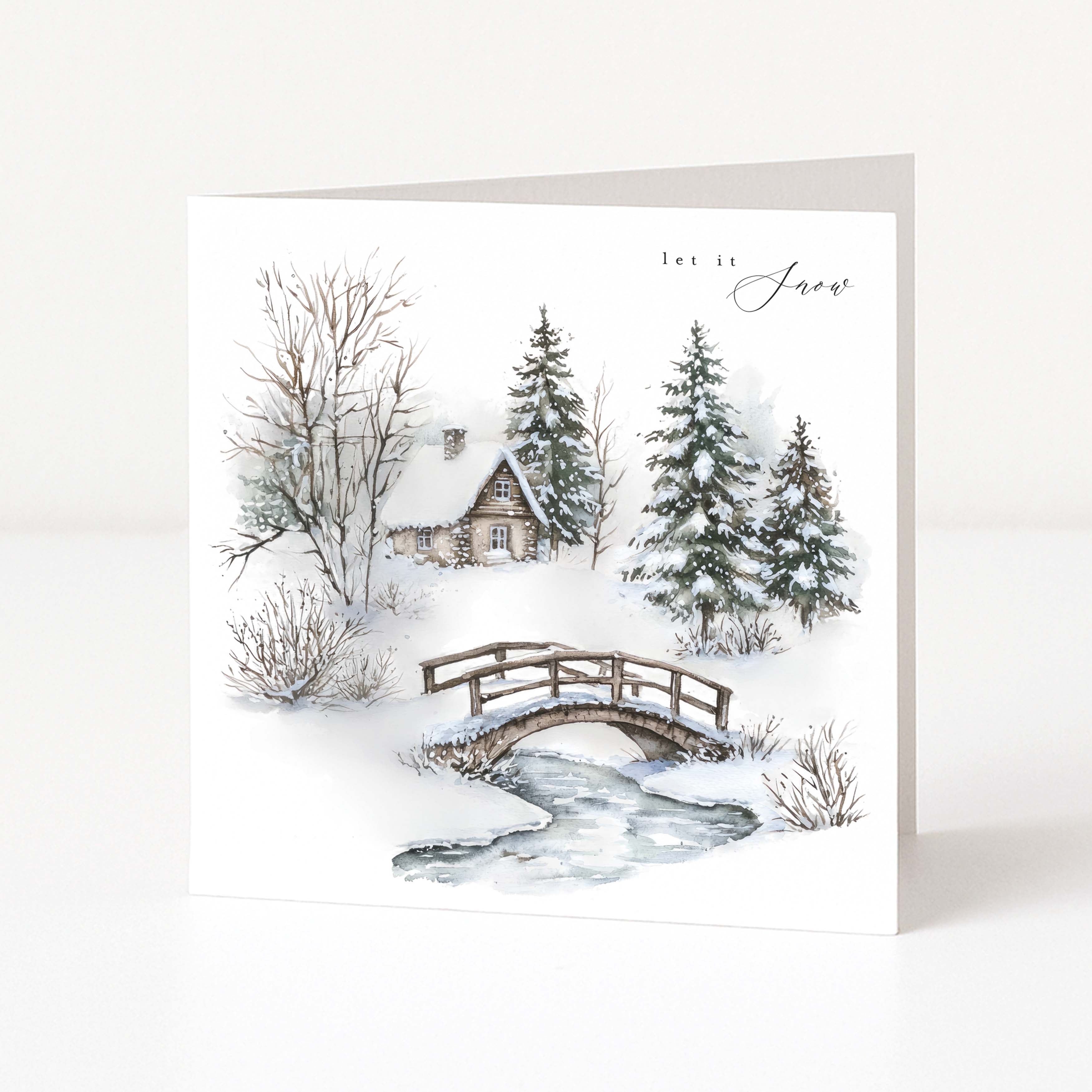A square Christmas greeting card standing upright on a white surface, featuring a winter landscape illustration with snow, evergreen trees, a small cabin, a wooden bridge, and a stream. The card has the words 'let it snow' printed at the top right.