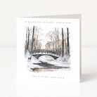A white Christmas greeting card standing upright on a white surface, featuring a winter landscape illustration with bare trees, snow, a small bridge, a deer, and the text 'WISHING YOU A MERRY CHRISTMAS' and 'AND A HAPPY NEW YEAR'.