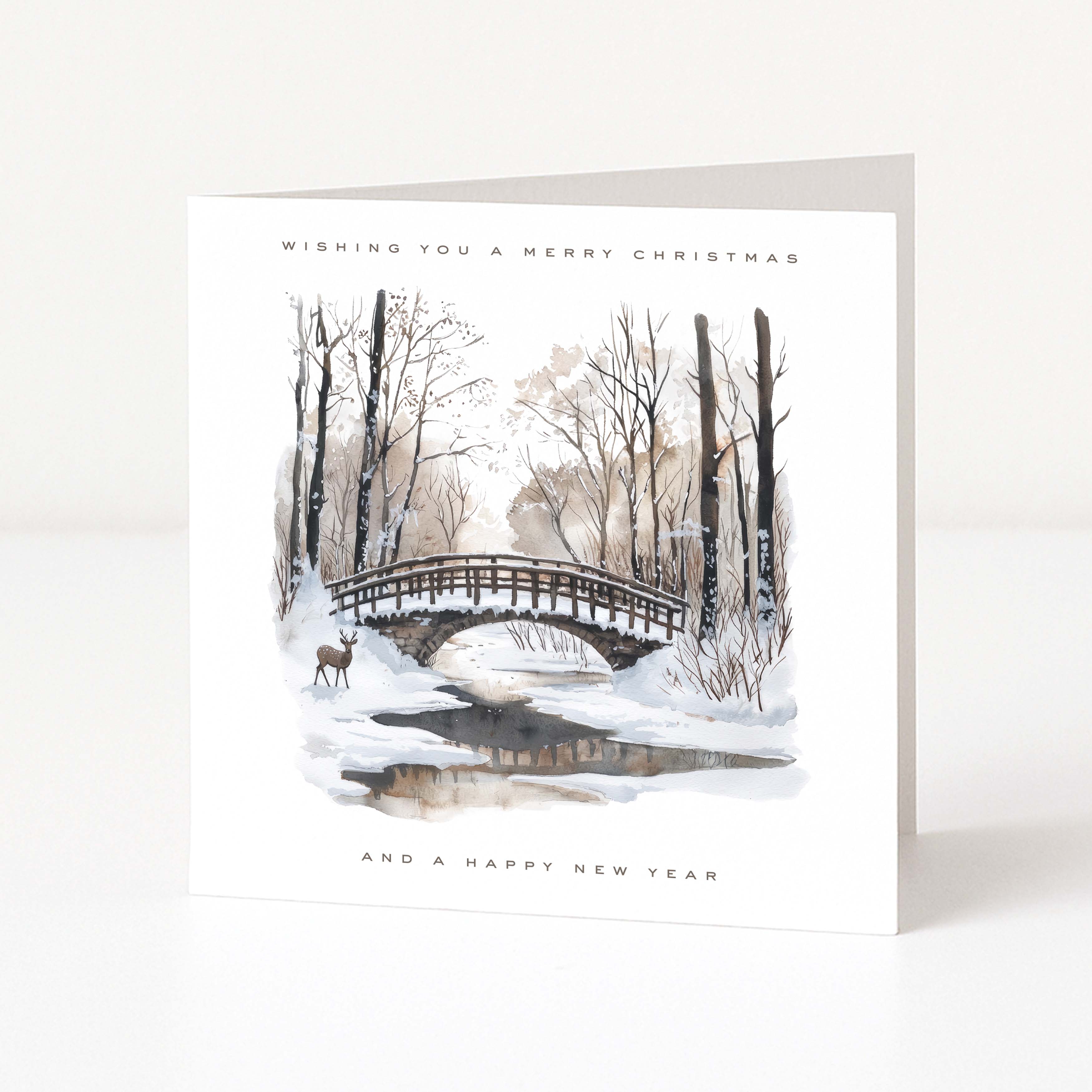 A white Christmas greeting card standing upright on a white surface, featuring a winter landscape illustration with bare trees, snow, a small bridge, a deer, and the text 'WISHING YOU A MERRY CHRISTMAS' and 'AND A HAPPY NEW YEAR'.