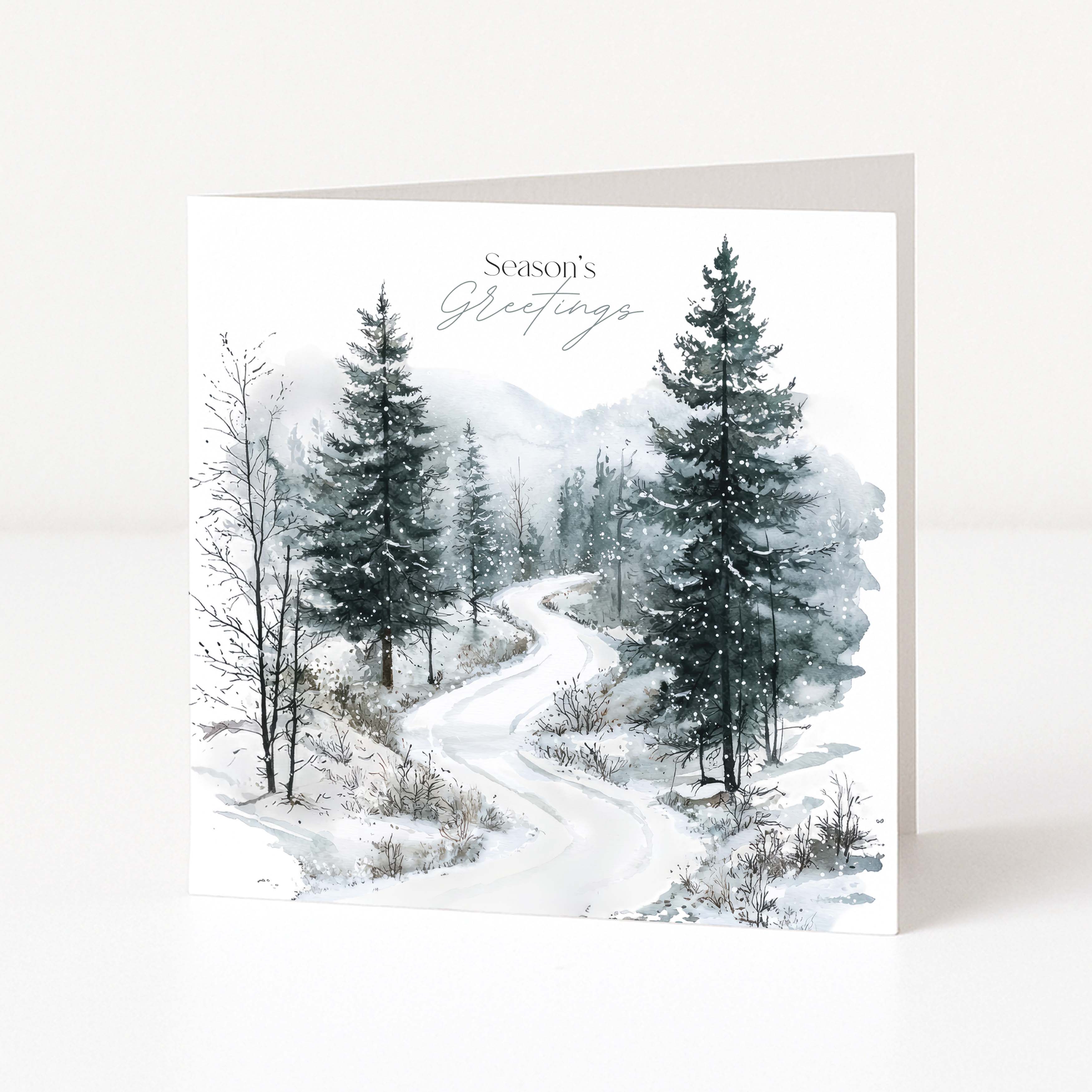 A square Christmas greeting card standing upright on a white surface, featuring a winter landscape illustration with snow-covered trees and a winding road. The card reads 'Season's Greetings' in elegant script near the top.