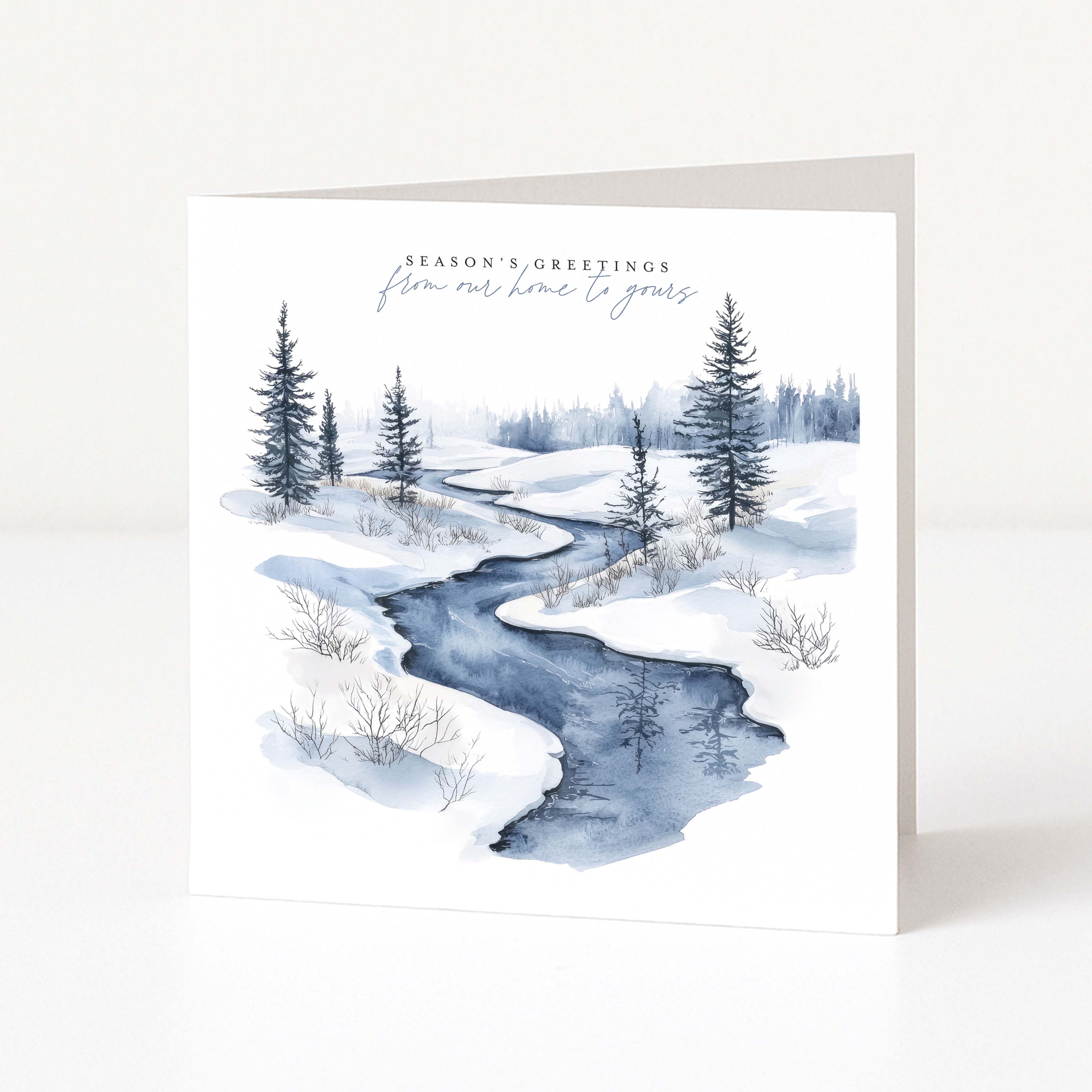 A square Christmas greeting card standing upright on a white surface, featuring a watercolor winter landscape with a winding blue stream, snow-covered ground, evergreen trees, and the text 'Season's Greetings' and 'from out of time to greet.'.