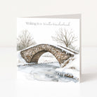A square Christmas greeting card standing upright on a white surface, featuring a watercolor winter scene with a stone bridge over a partially frozen stream, bare trees, and the text 'Walking in a Winter Wonderland' at the top.