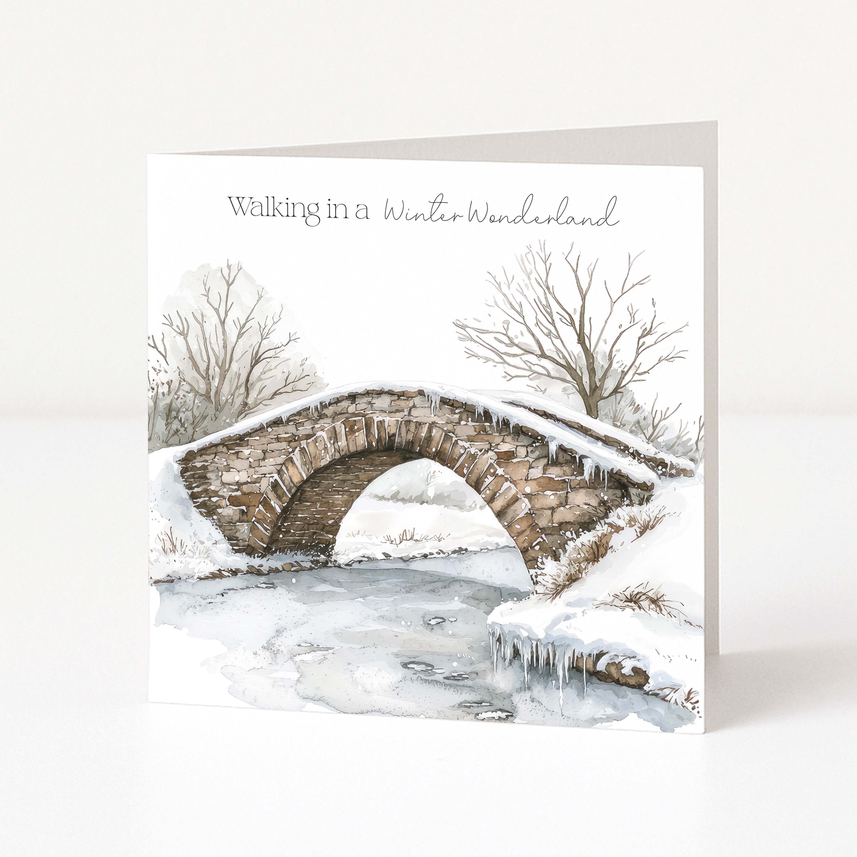 A square Christmas greeting card standing upright on a white surface, featuring a watercolor winter scene with a stone bridge over a partially frozen stream, bare trees, and the text 'Walking in a Winter Wonderland' at the top.